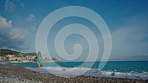 Happy young woman with backpack wearing in green blazer, denim shorts and sneakers walking along the sea on warm sunny