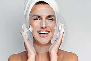 Happy young woman with bath towel on her head takes care of her skin face, applies cleansing foam after shower, smiling and