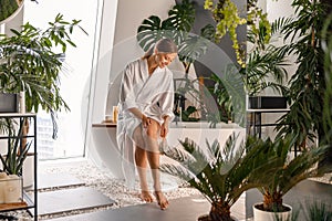 Happy young woman in bathrobe dry brushing her legs with a natural brush while sitting on the side of a bathtub in the