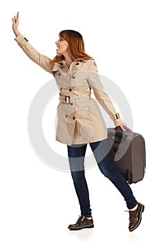Happy Young Woman In Beige Coat Is Standing With Suitcase, Holding Arm Raised And Waving