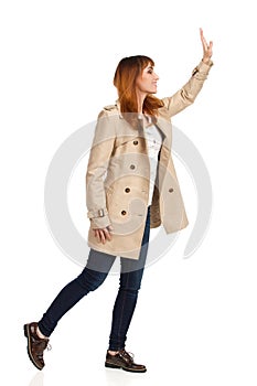 Happy Young Woman In Beige Coat Is Standing And Waving Hand