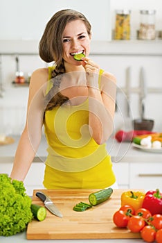 Happy young woman biting cucumber while cutting fresh salad