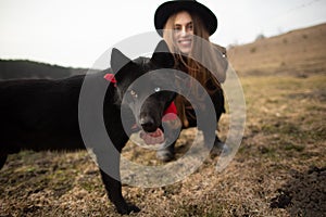 Happy young woman with black hat, plaing with her black dog on the shore of the lake
