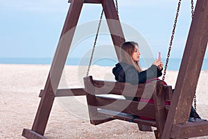 Happy young woman in black raincoat empty autumn sea beach. Smiling girl drinking tea with thermo can bottle. Lifestyle