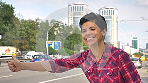 Happy young woman with black short hair lifting thumb and looking for taxi, standing on street near road in urban city