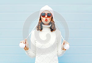 Happy young woman blowing red lips makes air kiss wearing a heart shape sunglasses, knitted hat, sweater over blue