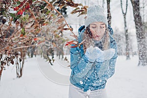 Happy young woman blows snow from hands having fun in winter forest. Girl wearing blue coat and mittens