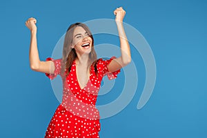 Happy young woman with bright smile in red casual dress looking up with excited face with hands up over blue background.