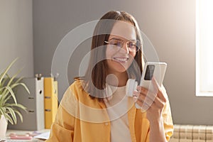 Happy young woman in casual clothes with a mobile phone in hands browsing smartphone, sitting in office at her workplace and