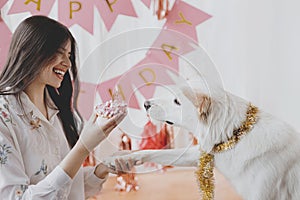 Happy young woman celebrating dog first birthday with donut and candle in room with pink decorations