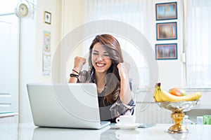 Happy young woman celebrating success with arms up in front of laptop at home.