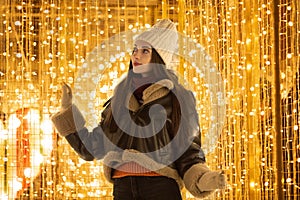 A happy young woman on Christmas night stands against the background of golden lights of garlands