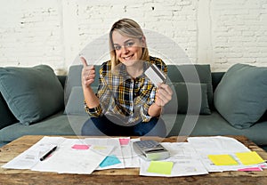 Happy young woman with credit card surrounded by papers calculating and paying bills