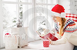 Happy young woman with cup of hot tea and book in winter window