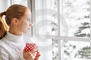 Happy young woman with cup of hot tea in winter window Christmas