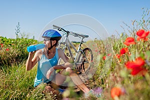 Happy young woman cyclist drinking water and having rest after riding bicycle in summer field.
