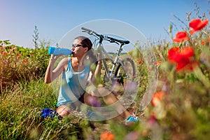 Happy young woman cyclist drinking water and having rest after riding bicycle in summer field among flowers. Sport