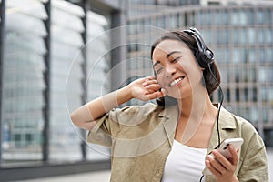 Happy young woman dancing on streets and listening music in headphones, holding smartphone