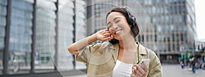 Happy young woman dancing on streets and listening music in headphones, holding smartphone