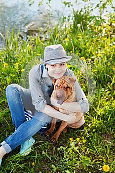 Happy young woman in denim overalls and hat hugging his beloved dog Shar Pei in the green grass in sunny day, true friends forever photo