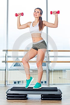 Happy young woman is doing exercises with dumbbells on step board.