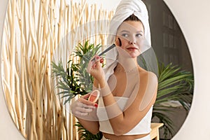 A happy young woman is doing makeup with a blush brush in front of the bathroom mirror.