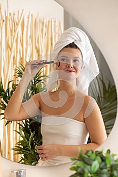 A happy young woman is doing makeup with a blush brush in front of the bathroom mirror.