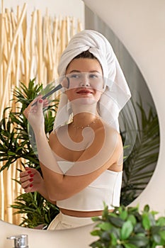 A happy young woman is doing makeup with a blush brush in front of the bathroom mirror.