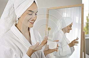 Happy young woman applying face cream while doing her skin care routine in the bathroom