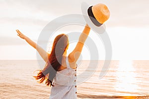 Happy young woman in a dress holding straw hat and and looking at the sunset on empty sand beach with her hands up. Freedoom,