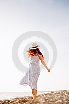 Happy young woman in dress and straw hat and walking alone on empty sand beach at sunset sea shore and smiling. Freedoom