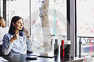 Happy young woman drinking cappuccino, latte, macchiato, tea, using tablet computer and talking on the phone in a coffee shop / ba