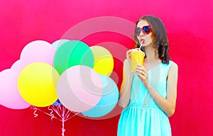 Happy young woman drinks a fruit juice from cup with an air colorful balloons over pink background