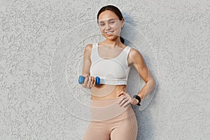 Happy young woman with dumbbell in hand making exercises outdoors in front of gray wall, looking smiling directly at camera,