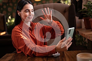 Happy young woman in earphones sitting at cafe making video call from her mobile phone. Caucasian girl at coffee shop