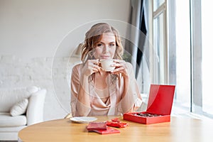 Happy young woman eating chocolate candies and drinking coffee