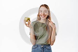 Happy young woman eating green apple, pointing at her white perfect smile, showing whitened healthy teeth, standing