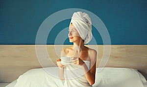 Happy young woman enjoying with cup of coffee drying her wet hair after bath with wrapped towel on her head sitting on bed looking