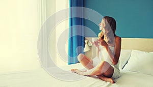 Happy young woman enjoying with cup of coffee sitting on bed looking out the window in the morning
