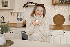 Happy young woman enjoying drinking tea or coffee in kitchen with Christmas decor over new year background