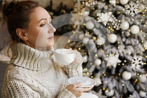 Happy young woman enjoying drinking tea or coffee in living room with Christmas decor