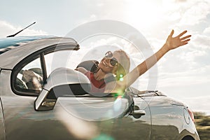 Happy young woman enjoying a ride in a convertible car. hand greeting. concept of road travel and adventure. riding with