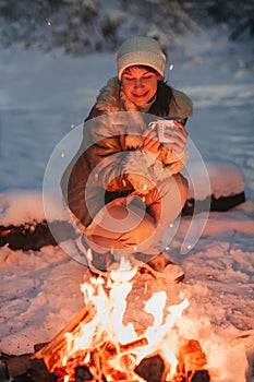 Happy young woman full of calmness and pacification in warm knitted hat and jacket sits on wooden sled by bonfire with cup