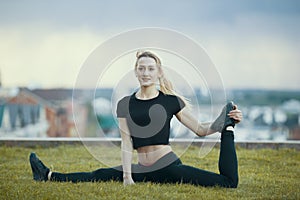 Happy young woman on the grass performs twine with the bent leg, on background cityscape