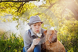 Happy young woman in a hat with dog Shar Pei sitting in the field in sunset light and blowing on a dandelion flowers