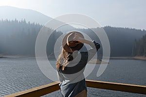 Happy young woman in hat enjoys lake view in mountains . Relaxing moments in forest. Freedom, people, lifestyle