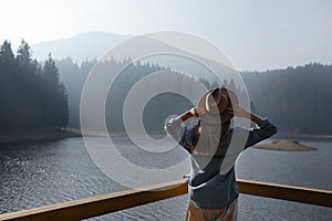 Happy young woman in hat enjoys lake view in mountains . Relaxing moments in forest. Freedom, people, lifestyle