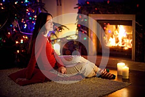 Happy young woman having a good time sitting by a fireplace in a cozy dark living room on Christmas eve. Celebrating Xmas at home