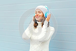 Happy young woman in headphones enjoys listens to music wearing a knitted hat, sweater over blue