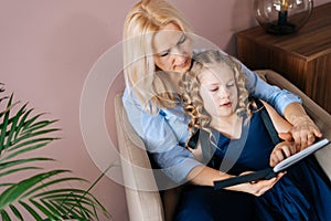 Happy young woman and her little curly daughter reading book together while sitting on armchair